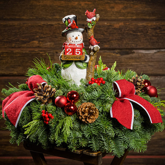 A centerpiece of noble fir, cedar, and pine with red ball ornaments, faux red berries, pinecones, 3 red bow tucks, and a decoration of a resin snowman holding 2 red blocks with numbers representing the number of days left until Christmas. Next to the black-top hat-wearing snowman is a frosted tree with 2 red cardinals.