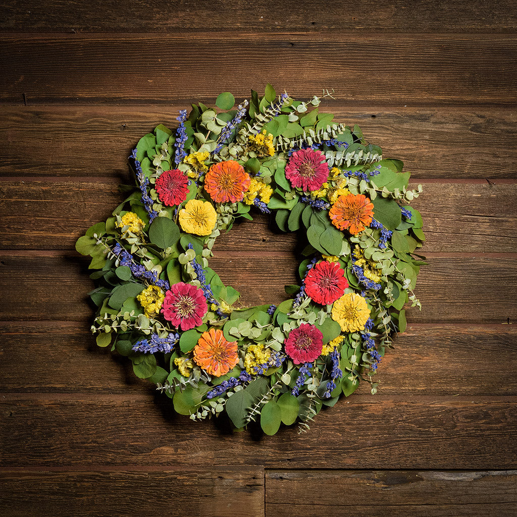 A dried wreath made of green silver dollar eucalyptus leaves, green spiral eucalyptus, yellow sinuata flowers, purple larkspur, naturally dried and naturally preserved zinnias in yellow, fuchsia pink and orange hung on a dark wood background.