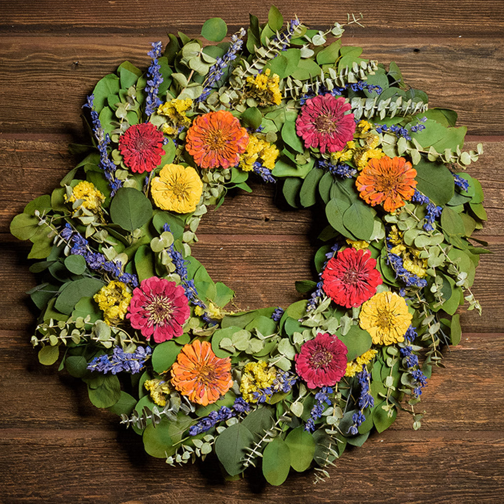 A close-up of a dried wreath made of green silver dollar eucalyptus leaves, green spiral eucalyptus, yellow sinuata flowers, purple larkspur, naturally dried and naturally preserved zinnias in yellow, fuchsia pink and orange hung on a dark wood background.