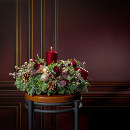 A festive Christmas wreath centerpiece displayed on a small wooden stool against a dark burgundy paneled wall. The wreath is decorated with green pine branches, red berries, pinecones, gold ornaments, and two red pillar candles.