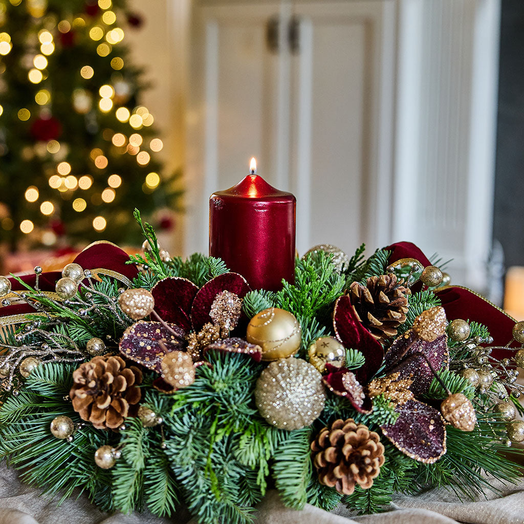 A festive Christmas wreath centerpiece with a lit dark red pillar candle, green pine branches, gold and red ornaments, pinecones, and ribbon, set indoors with a blurred Christmas tree in the background.