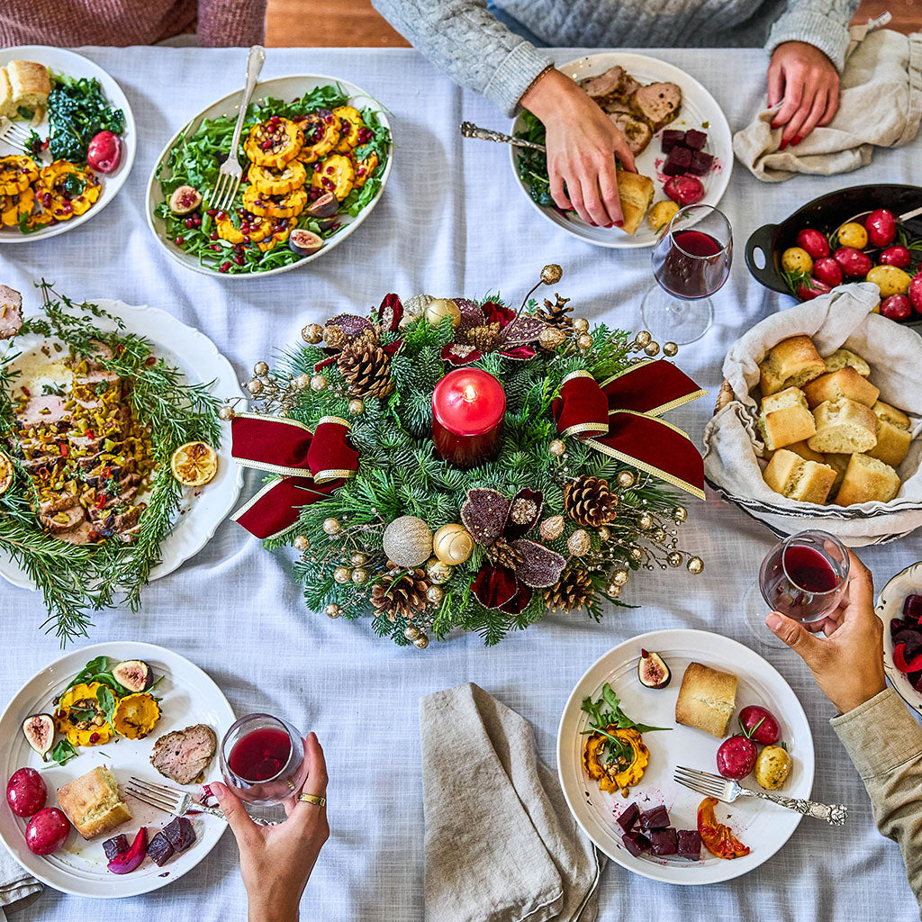 Overhead view of a holiday dining table set with plates of food, bread, red wine, and a festive green wreath centerpiece with red ribbons and a candle, with hands reaching in from around the table.