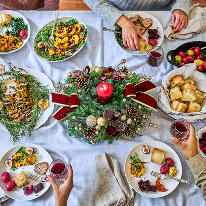 Overhead view of a holiday dining table set with plates of food, bread, red wine, and a festive green wreath centerpiece with red ribbons and a candle, with hands reaching in from around the table.