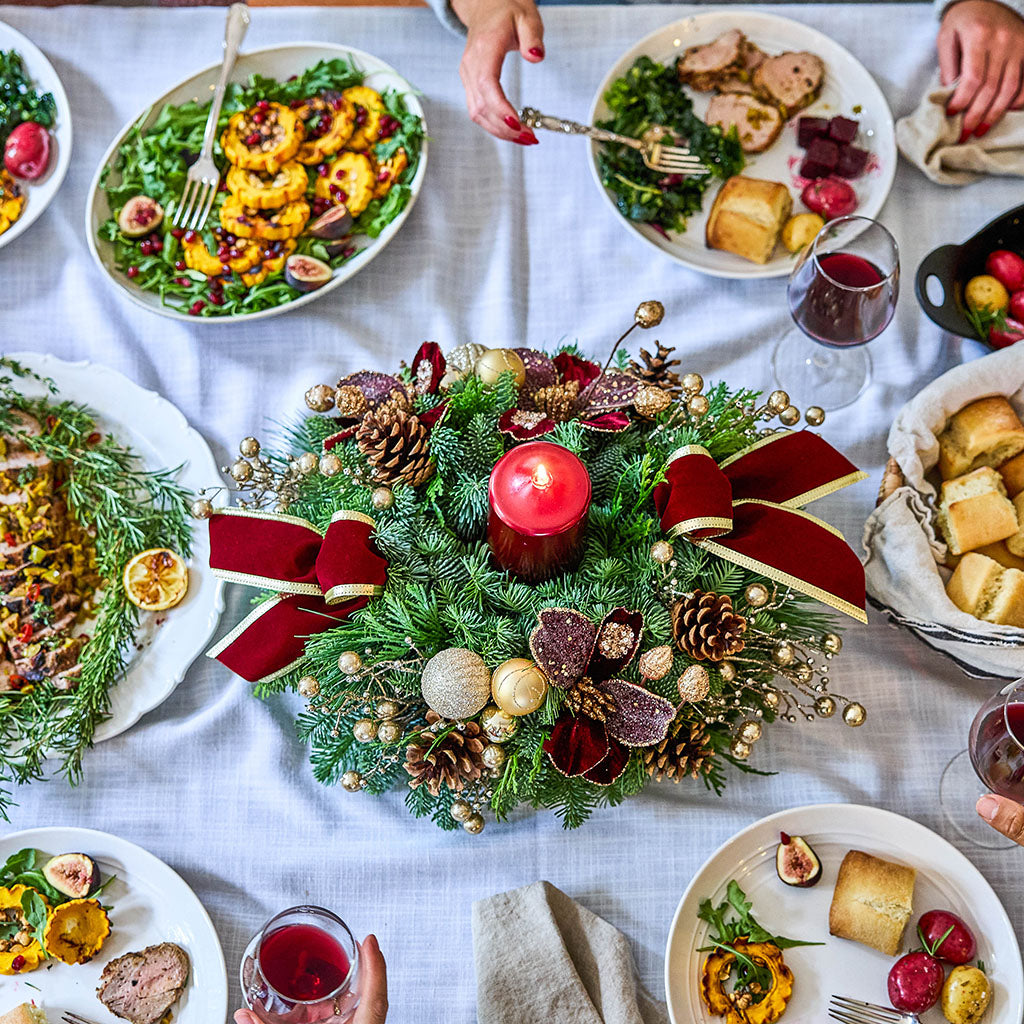 A festive Christmas wreath centerpiece on a white tablecloth, surrounded by a holiday table setting with plates of food, red wine, and hands reaching in from the top and bottom edges.