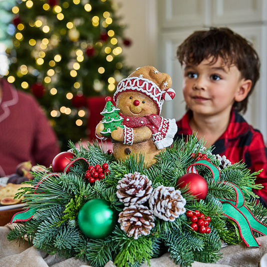 A festive Christmas wreath decoration featuring a gingerbread man ornament, green pine branches, red ornaments, pinecones, and ribbons, with a young boy and another person blurred in the background in front of a lit Christmas tree.
