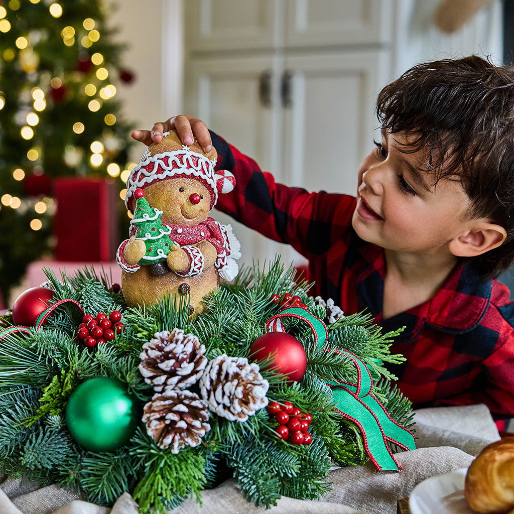 A young boy in a red and black plaid shirt reaches toward a gingerbread teddy bear ornament hanging above a decorated Christmas wreath with pinecones, green ornaments, and red berries in a cozy indoor holiday setting.