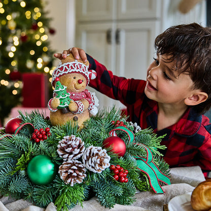 A young boy in a red and black plaid shirt reaches toward a gingerbread teddy bear ornament hanging above a decorated Christmas wreath with pinecones, green ornaments, and red berries in a cozy indoor holiday setting.