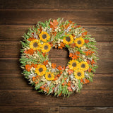 A dried wreath made of green oats with gray sage, cream globe amaranthus, flax pods, orange preserved oak leaves, and preserved sunflowers hung on a dark wood background.