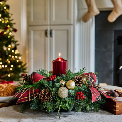 A festive Christmas wreath centerpiece on a table, decorated with green pine branches, red plaid ribbons, pine cones, gold ornaments, red berries, and a lit red pillar candle. The background shows a softly blurred holiday interior with a Christmas tree, fireplace, and stockings.