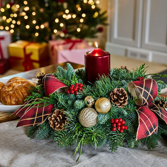 A festive Christmas centerpiece with a green pine wreath, red pillar candle, red plaid ribbon bows, pinecones, gold ornaments, and red berries on a table, with a blurred Christmas tree and wrapped gifts in the background.
