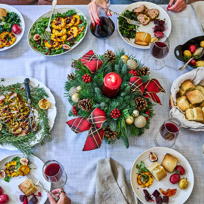 Overhead view of a festive holiday table setting with a central green pine wreath centerpiece decorated with red plaid bows, pinecones, gold ornaments, and a red pillar candle. Plates of food, glasses of red wine, and hands dining are visible around the table.