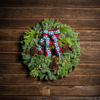 An evergreen wreath of noble fir, cedar, and juniper with natural pinecones, faux red berries, and a red brushed-linen &amp; blue starred bow on a dark wood background.