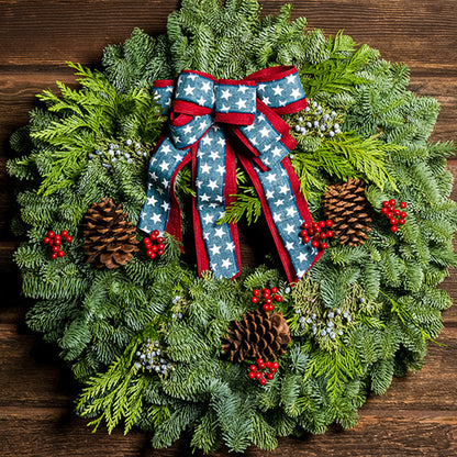 An evergreen wreath of noble fir, cedar, and juniper with natural pinecones, faux red berries, and a red brushed-linen &amp; blue starred bow on a dark wood background.