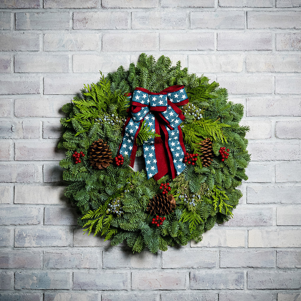 An evergreen wreath of noble fir, cedar, and juniper with natural pinecones, faux red berries, and a red brushed-linen &amp; blue starred bow on a white brick background.