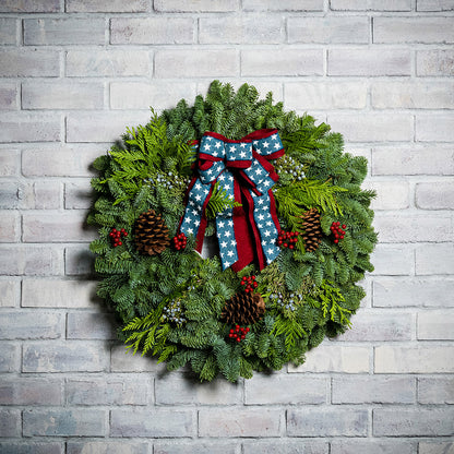 An evergreen wreath of noble fir, cedar, and juniper with natural pinecones, faux red berries, and a red brushed-linen &amp; blue starred bow on a white brick background.