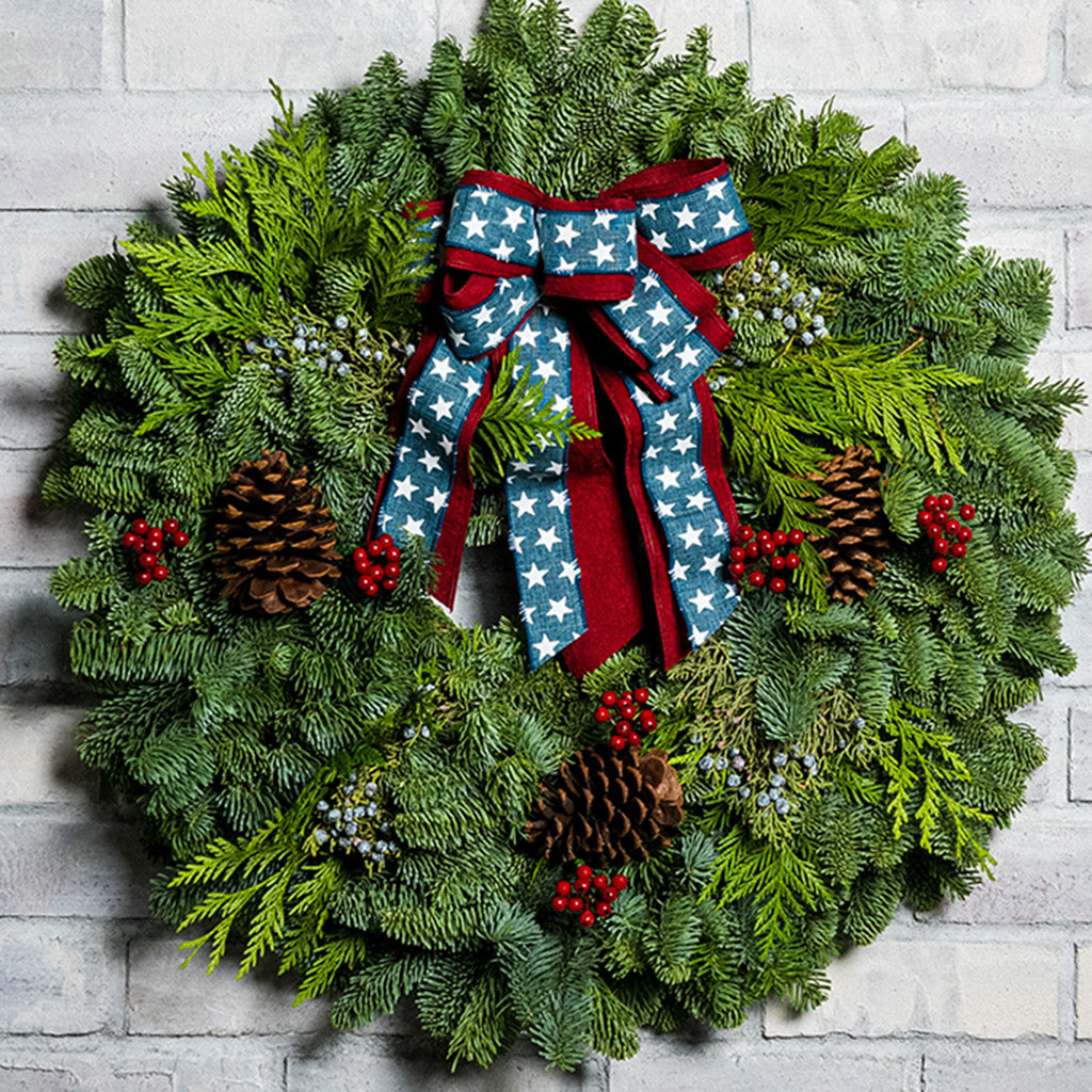 An evergreen wreath of noble fir, cedar, and juniper with natural pinecones, faux red berries, and a red brushed-linen &amp; blue starred bow on a white brick background.