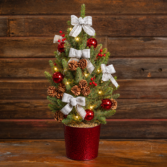 A small artificial Christmas tree in a red textured pot, decorated with white plaid bows, red ornaments, pinecones, and warm white string lights, set against a dark wood plank background.