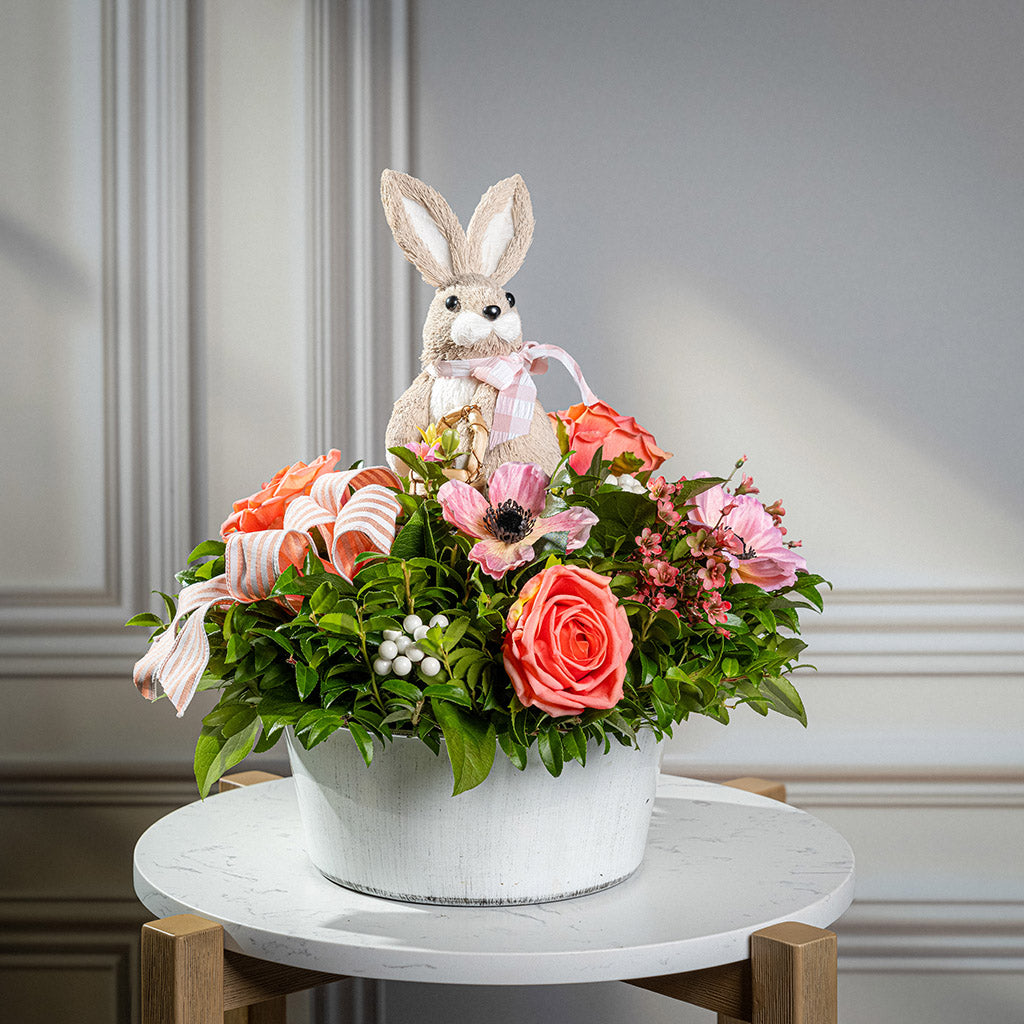 Fresh salal and green huckleberry with faux peachy-orange roses, faux pink anemone flowers, faux pink wax flowers, faux white berries, a peach striped bow, and a natural sisal rabbit in a white metal pot