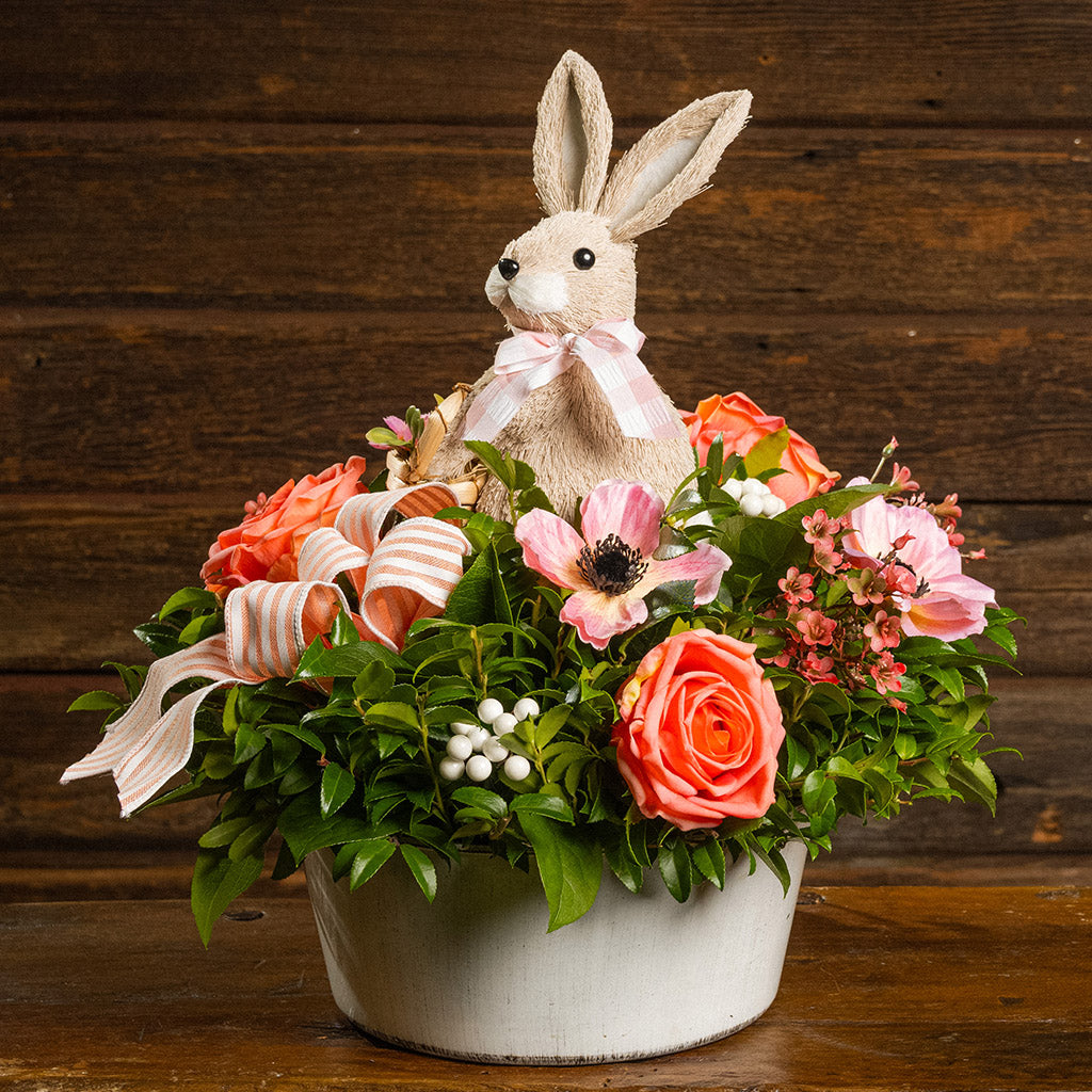 Fresh salal and green huckleberry with faux peachy-orange roses, faux pink anemone flowers, faux pink wax flowers, faux white berries, a peach striped bow, and a natural sisal rabbit in a white metal pot