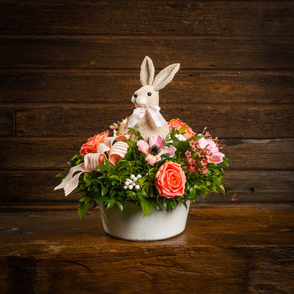 Fresh salal and green huckleberry with faux peachy-orange roses, faux pink anemone flowers, faux pink wax flowers, faux white berries, a peach striped bow, and a natural sisal rabbit in a white metal pot