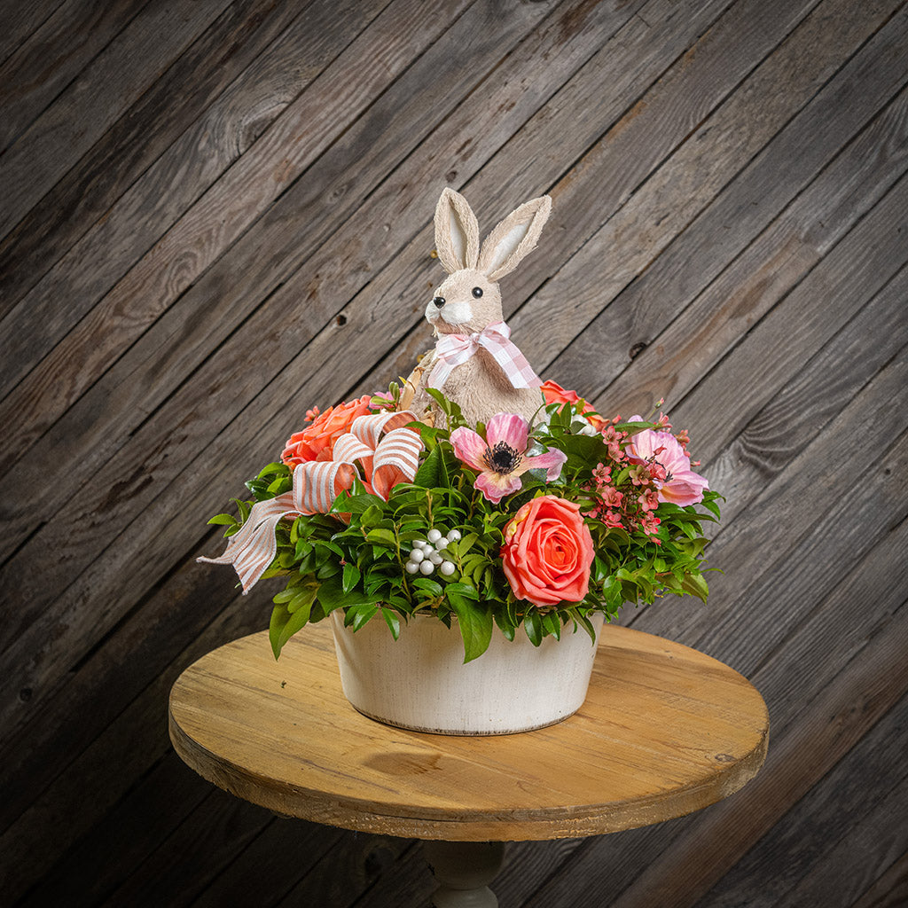 Fresh salal and green huckleberry with faux peachy-orange roses, faux pink anemone flowers, faux pink wax flowers, faux white berries, a peach striped bow, and a natural sisal rabbit in a white metal pot