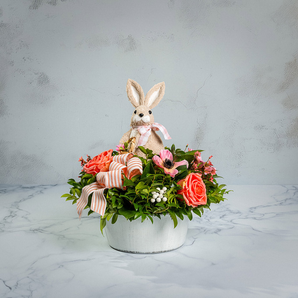 Fresh salal and green huckleberry with faux peachy-orange roses, faux pink anemone flowers, faux pink wax flowers, faux white berries, a peach striped bow, and a natural sisal rabbit in a white metal pot