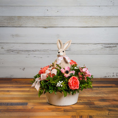 Fresh salal and green huckleberry with faux peachy-orange roses, faux pink anemone flowers, faux pink wax flowers, faux white berries, a peach striped bow, and a natural sisal rabbit in a white metal pot