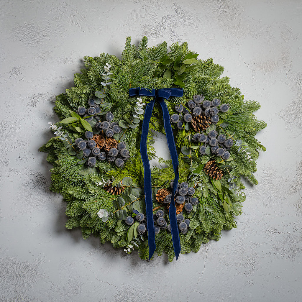 Christmas wreath with greenery, pinecones, and a blue ribbon on a light gray background