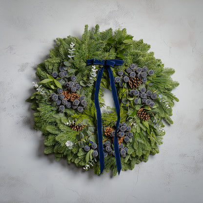 Christmas wreath with greenery, pinecones, and a blue ribbon on a light gray background