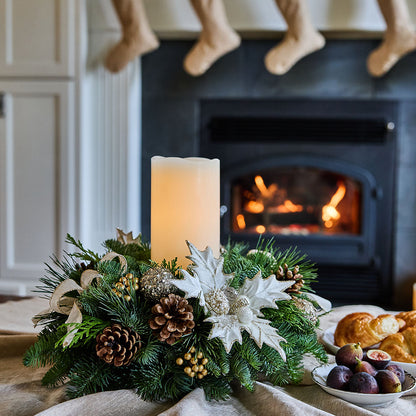 A festive Christmas wreath centerpiece with pine branches, white poinsettias, pinecones, and a tall white pillar candle, styled on a table in front of a lit fireplace with stockings hanging from the mantel and plates of pastries and figs nearby.