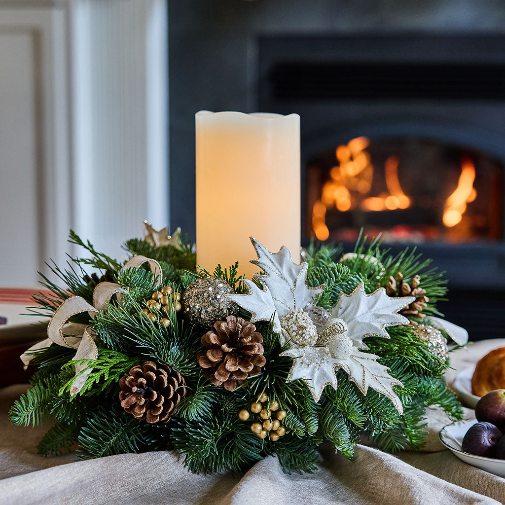 A festive Christmas wreath centerpiece with a lit cream pillar candle, green pine branches, brown pinecones, white poinsettia-style flowers, and gold berry accents, set on a table with a blurred fireplace in the background.