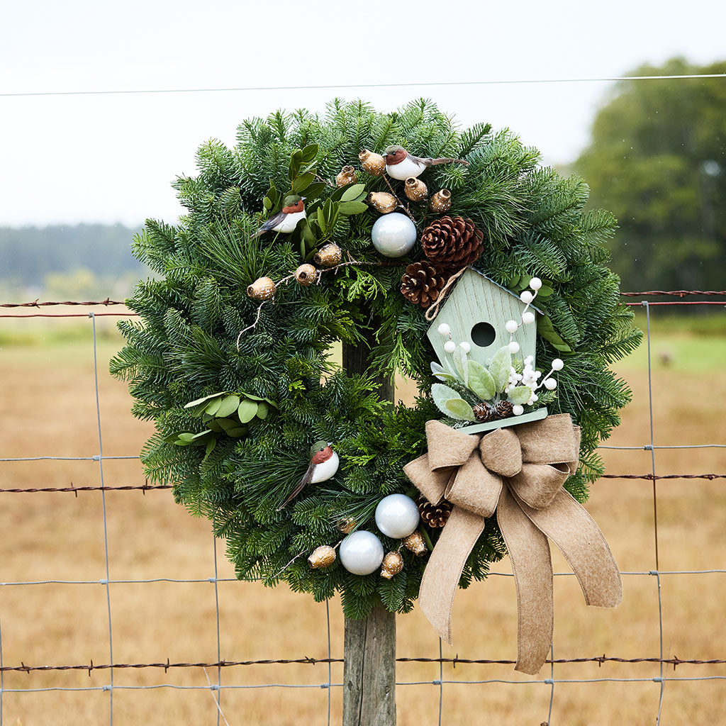 A festive evergreen wreath hanging on a wire fence, decorated with a light green birdhouse ornament, a large tan burlap bow, white and gold baubles, pinecones, and small bird figurines, with a blurred field and trees in the background.