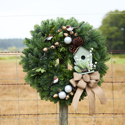 A festive evergreen wreath hanging on a wire fence, decorated with a light green birdhouse ornament, a large tan burlap bow, white and gold baubles, pinecones, and small bird figurines, with a blurred field and trees in the background.