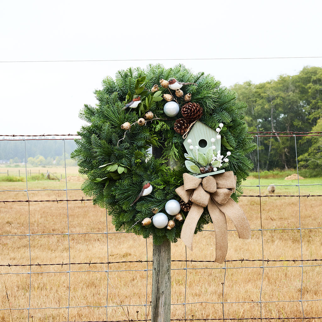 A green pine wreath decorated with a white birdhouse, burlap bow, white eggs, pinecones, and small bird figurines, hanging on a wire fence in a rural field.