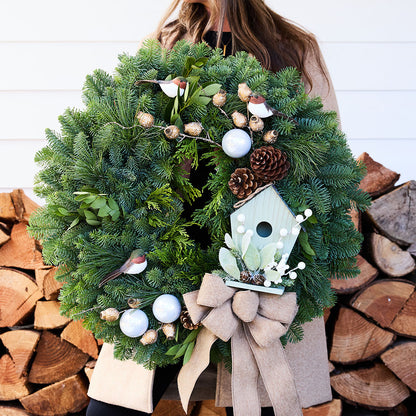 A person holds a large green evergreen wreath decorated with pinecones, white ornaments, small bird figurines, and a light blue birdhouse with a beige ribbon bow. The background shows stacked firewood and a white paneled wall.