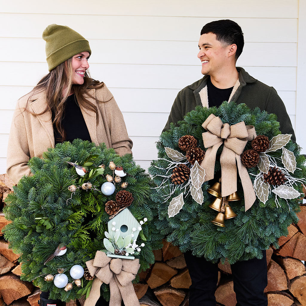 A man and a woman standing side by side holding decorated Christmas wreaths in front of a white wood-paneled wall and stacked firewood. The woman wears an olive green beanie and tan coat, and the man wears an olive green hoodie.