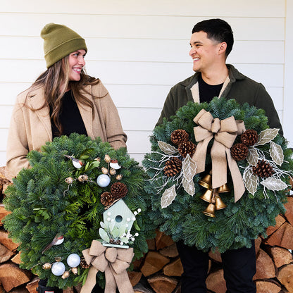 A man and a woman standing side by side holding decorated Christmas wreaths in front of a white wood-paneled wall and stacked firewood. The woman wears an olive green beanie and tan coat, and the man wears an olive green hoodie.