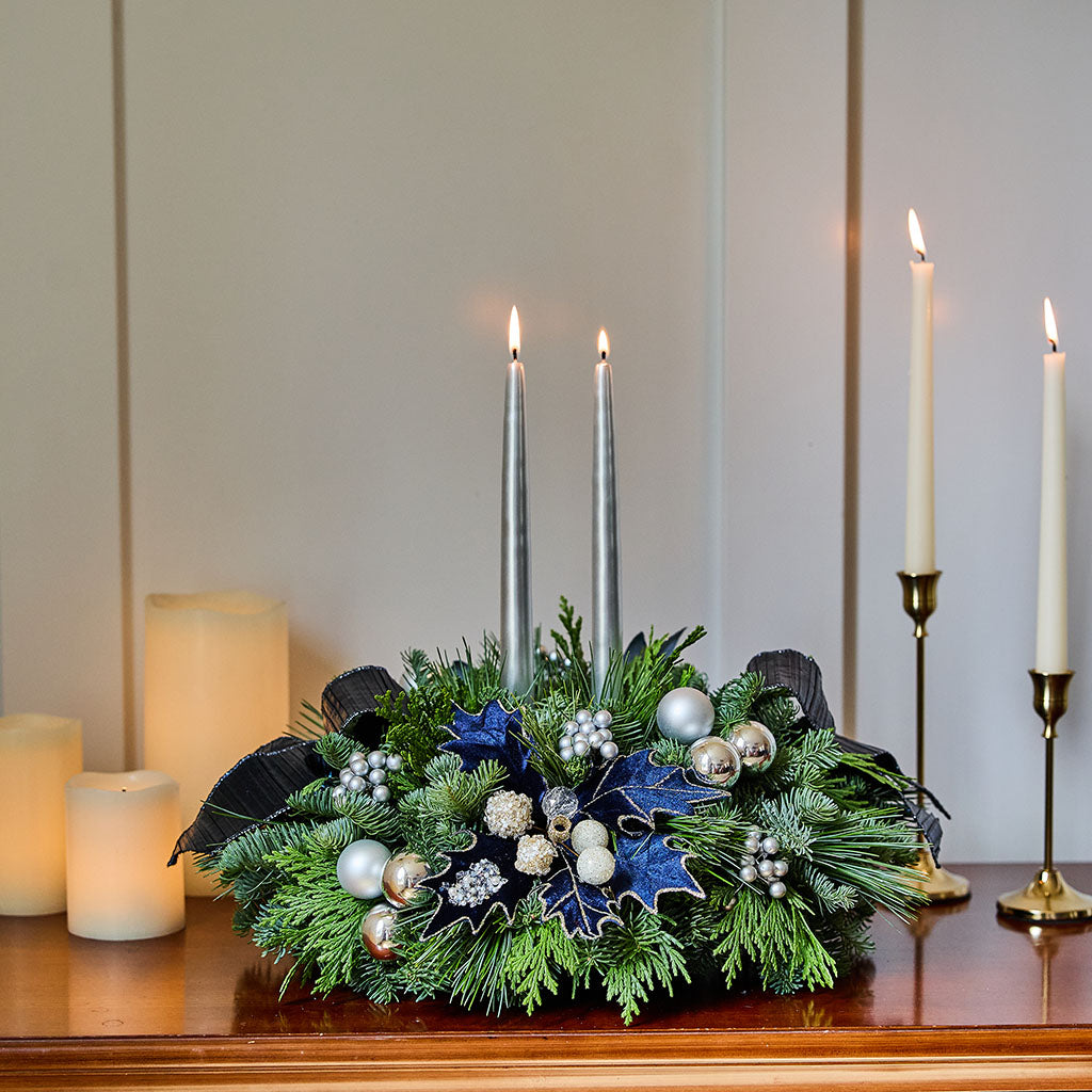 A festive Christmas wreath centerpiece with green pine branches, blue holly leaves, silver and white ornaments, and two tall silver taper candles, displayed on a wooden mantel with lit pillar candles in the background.