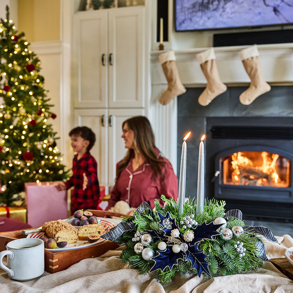 A cozy holiday living room scene with a woman and child near a fireplace and Christmas tree, with a decorated green wreath and lit candles in the foreground.