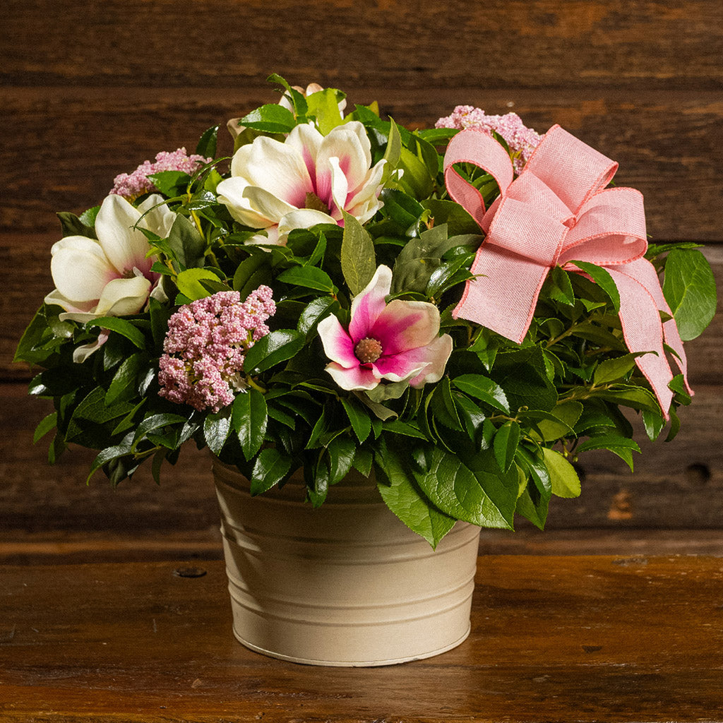 Fresh salal and green huckleberry with faux magnolia flowers, faux pink verbena, and a mauve pink bow in a white metal pot