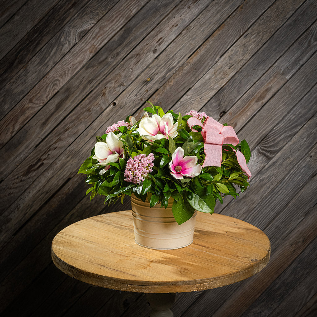 Fresh salal and green huckleberry with faux magnolia flowers, faux pink verbena, and a mauve pink bow in a white metal pot