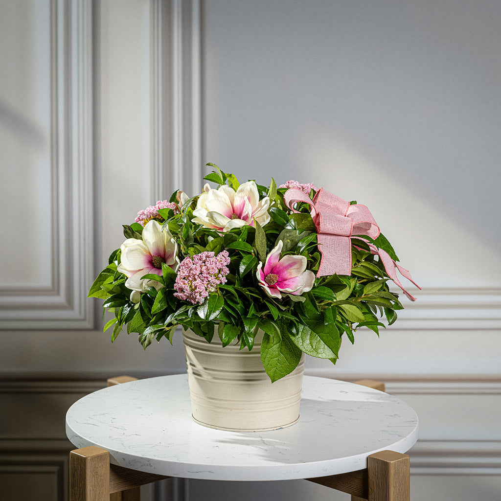 Fresh salal and green huckleberry with faux magnolia flowers, faux pink verbena, and a mauve pink bow in a white metal pot