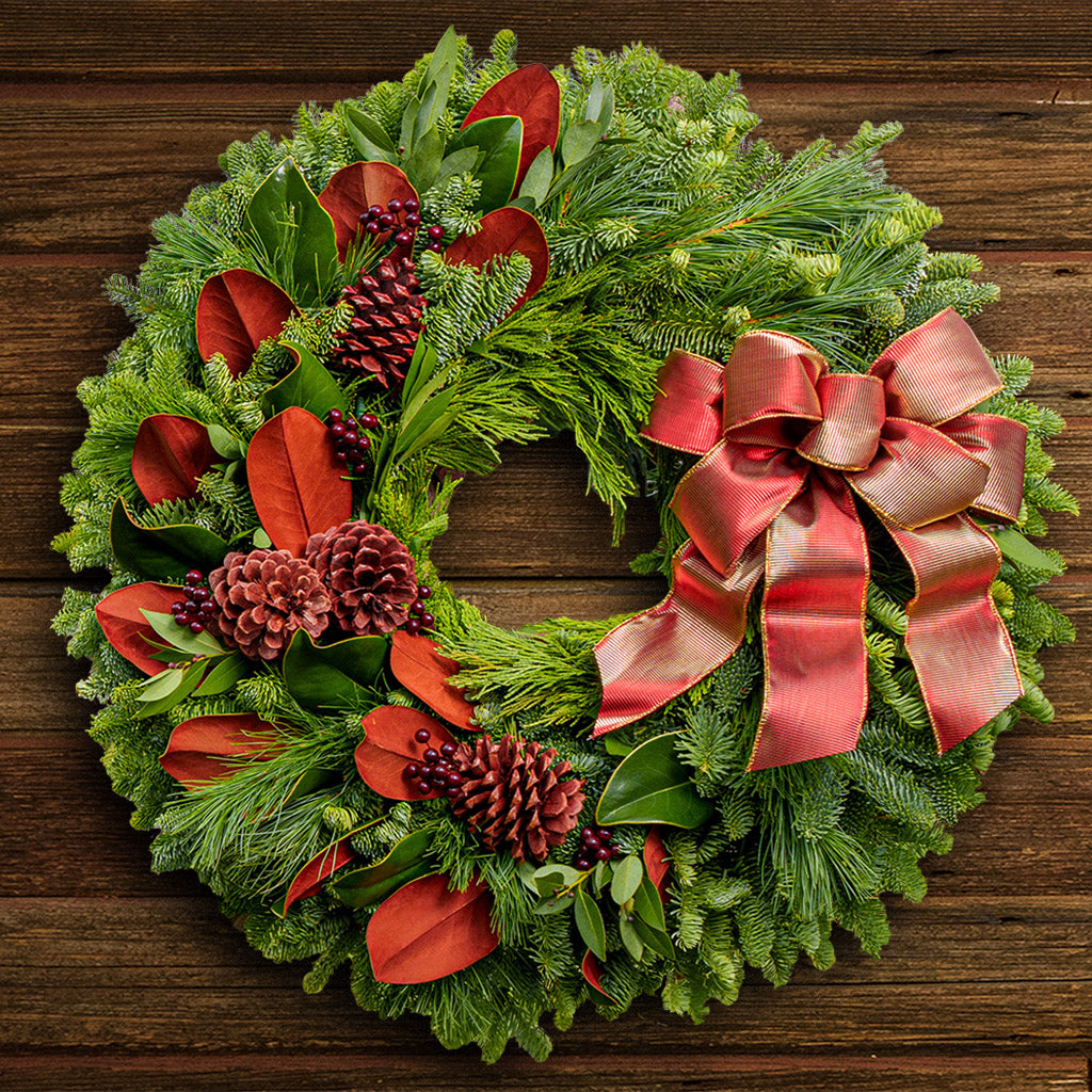 A wreath made of noble fir, magnolia leaves, bay leaves, white pine and incense cedar with ponderosa pine cones, faux burgundy berry clusters, and a shimmery gold/reddish-orange bow