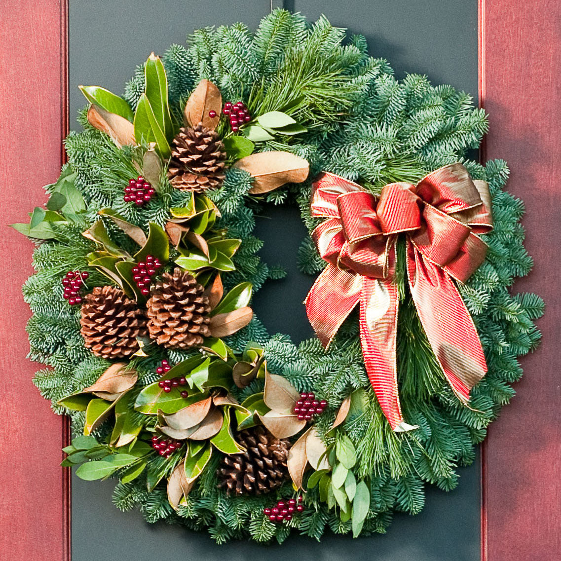A wreath made of noble fir, magnolia leaves, bay leaves, white pine and incense cedar with ponderosa pine cones, faux burgundy berry clusters, and a shimmery gold/reddish-orange bow hung on a black and red door.