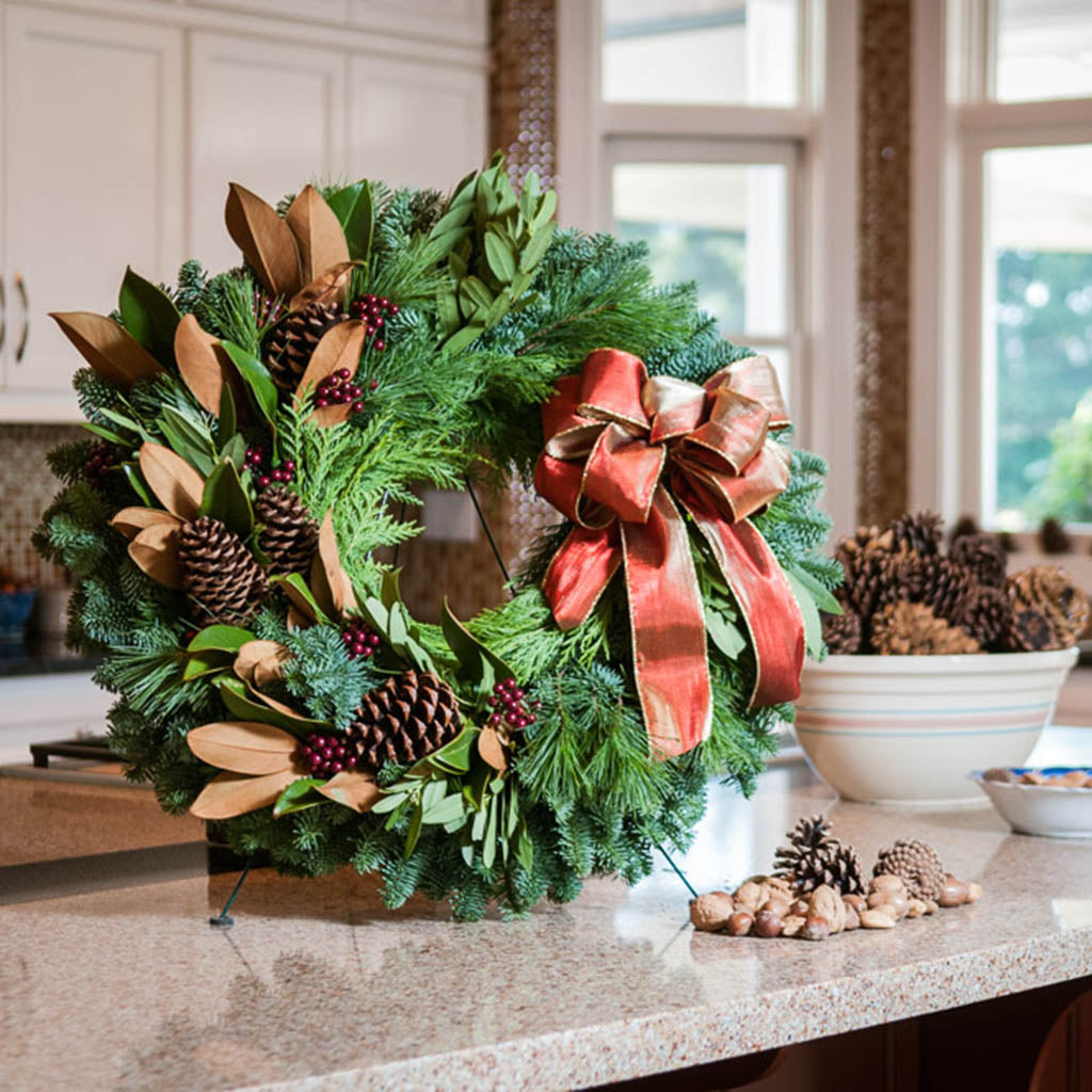 A wreath made of noble fir, magnolia leaves, bay leaves, white pine, and incense cedar with ponderosa pine cones, faux burgundy berry clusters, and a shimmery gold/reddish-orange sitting on a wreath easel on a countertop. 
