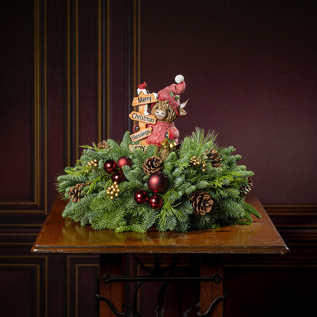 Decorative Christmas wreath with ornaments and a small figure on a wooden table against a dark background.