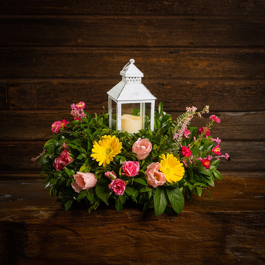 Fresh salal and green huckleberry with faux yellow gerbera daisies, faux mini pink roses, a mix of mini faux pink flowers, and faux tulips with a white LED lantern