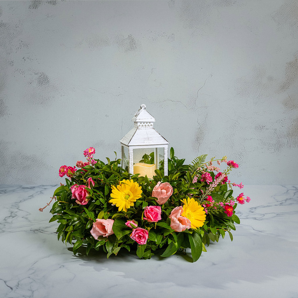 Fresh salal and green huckleberry with faux yellow gerbera daisies, faux mini pink roses, a mix of mini faux pink flowers, and faux tulips with a white LED lantern