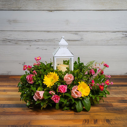 Fresh salal and green huckleberry with faux yellow gerbera daisies, faux mini pink roses, a mix of mini faux pink flowers, and faux tulips with a white LED lantern