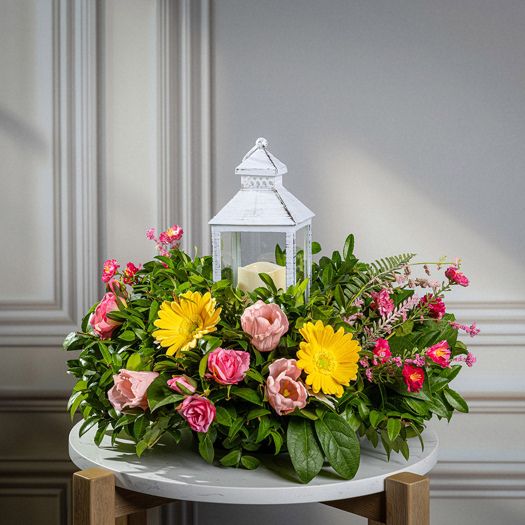 Fresh salal and green huckleberry with faux yellow gerbera daisies, faux mini pink roses, a mix of mini faux pink flowers, and faux tulips with a white LED lantern